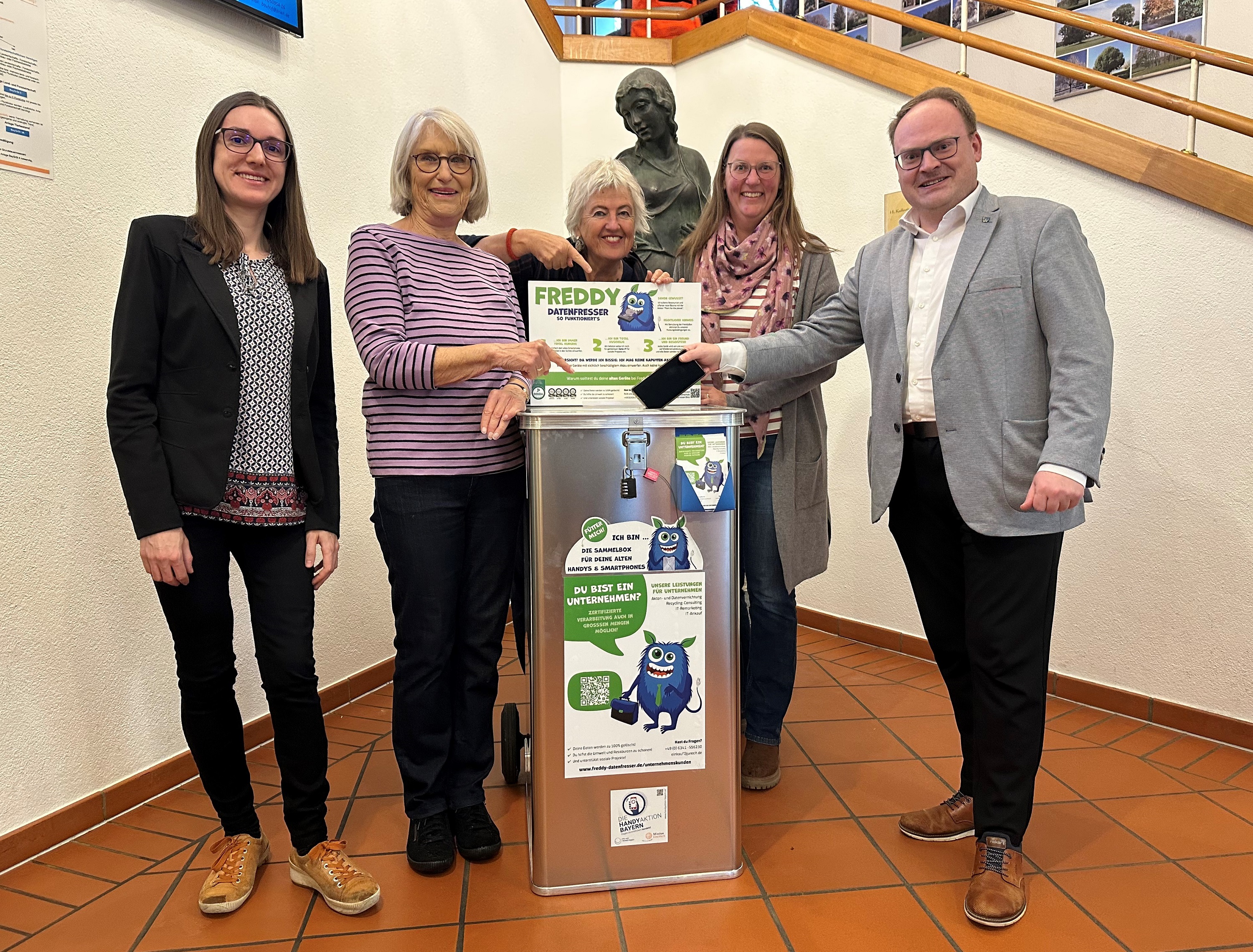 Im Eingangsbereich des Rathauses ist das Priener Sammelzentrum der „HandyAktion Bayern“. (v.l.n.r.) Die Büchereileiterin Melanie Schieber, die Fairtrade-Beauftragte und Gemeinderätin Angela Kind, die Inhaberin des Eine-Welt-Ladens Monica Lieschke, die Fairtrade-Beauftragte und Projektkoordinatorin Bettina Bartsch und der Erste Bürgermeister der Marktgemeinde Andreas Friedrich füttern die Freddy-Alu-Sammelbox mit ihren Althandys. 
Bildrechte: Markt Prien a. Chiemsee